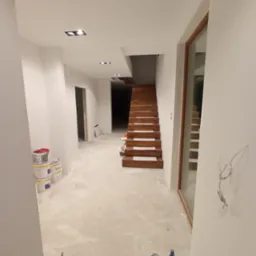 Wnętrze domu w Gnieznie po malowaniu ścian na biało, z widocznymi schodami i oknem. Ślady farby na podłodze sugerują trwające prace wykończeniowe.