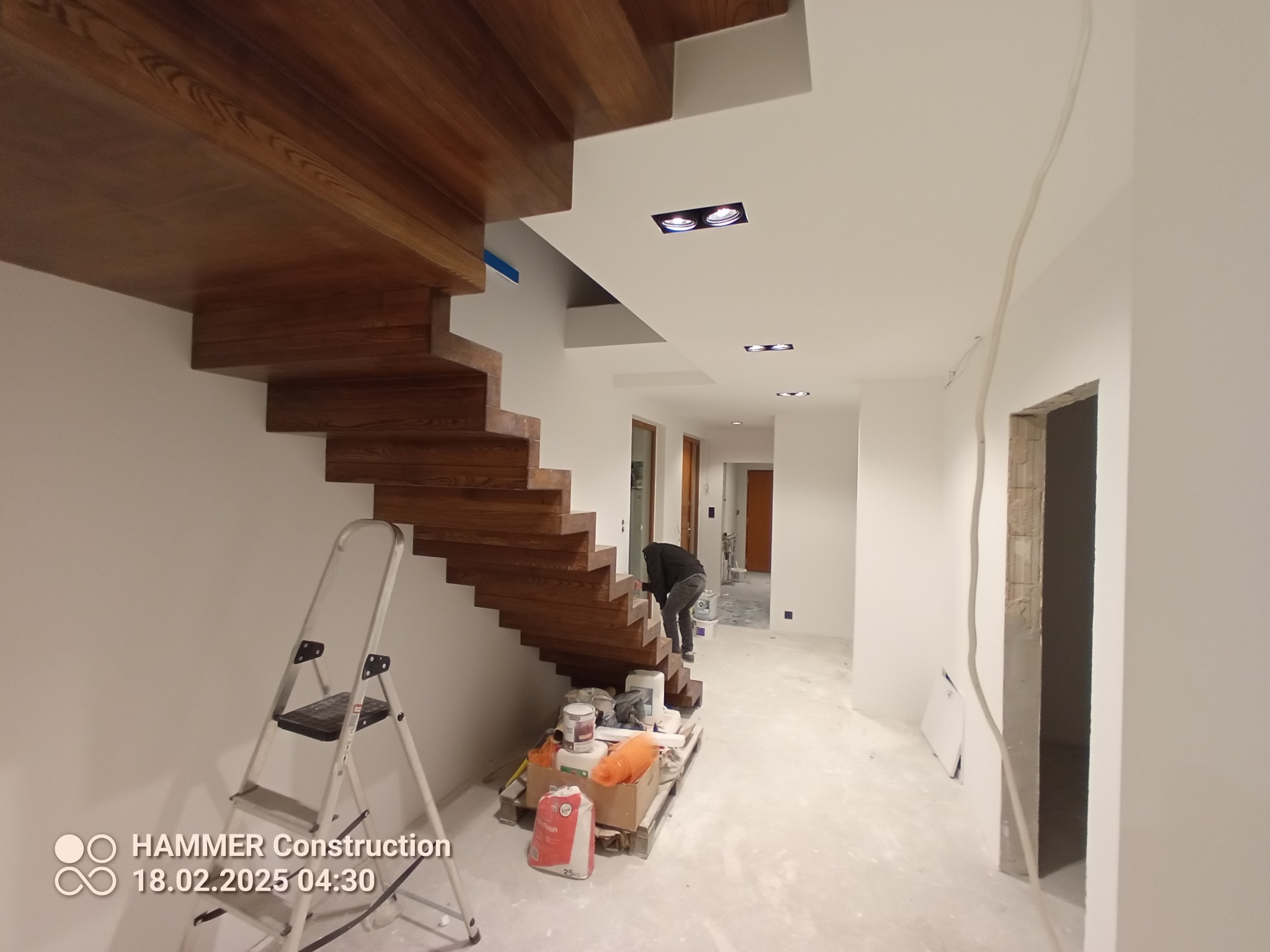 Wnętrze w trakcie wykańczania: drewniane schody, drabina malarska, materiały budowlane i pracownik przy ścianie. Widoczne instalacje elektryczne.