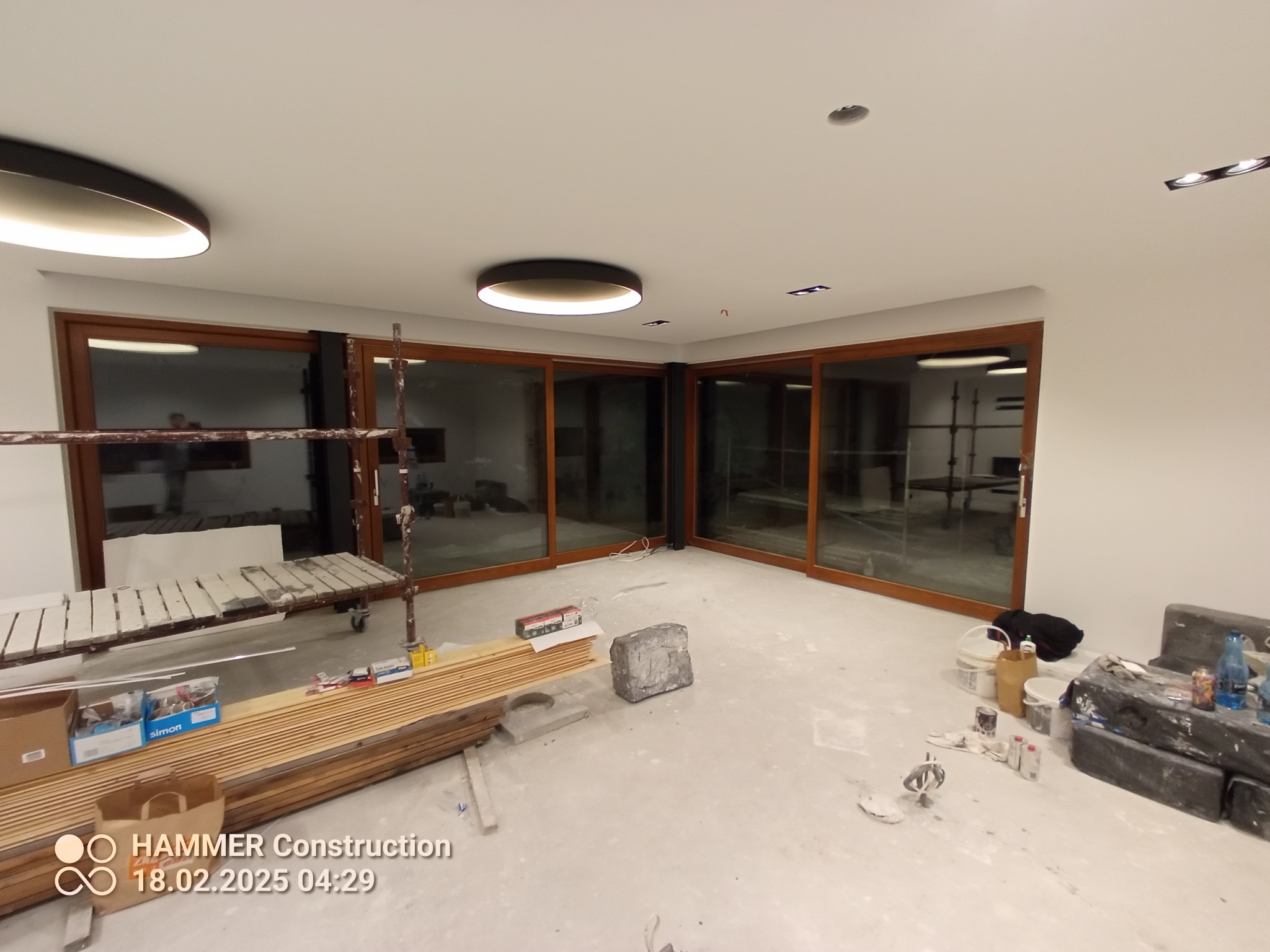 Wnętrze w trakcie remontu: widoczne okna przesuwne z drewnianymi ramami, rusztowanie, materiały budowlane i narzędzia. Na suficie zamontowane nowoczesne lampy.