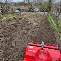 MM GardenTech Mateusz Gałek - Czerwony glebogryzarek EcoAgrino 750P przygotowuje ziemię pod uprawę w ogrodzie, widoczny fragment ogrodu z ogrodzeniem oraz osoba w tle.