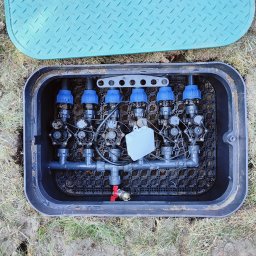 MM GardenTech Mateusz Gałek - Otwarta skrzynka z zaworami i rurami systemu nawadniania, widoczna zielona pokrywa obok.