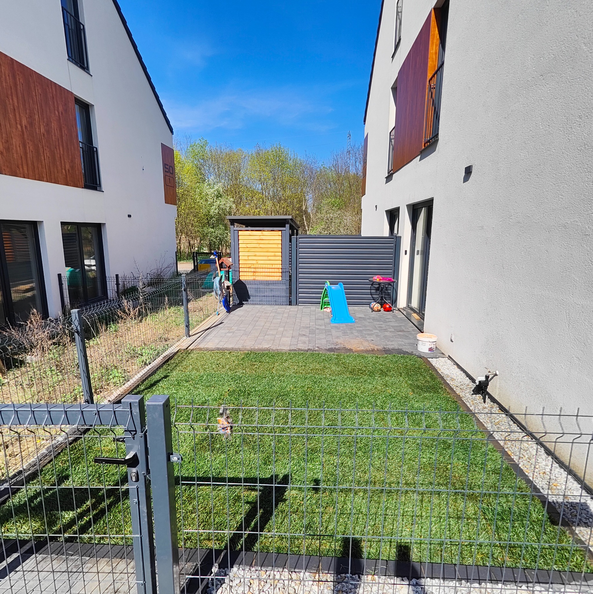 Niewielki ogród z trawnikiem z rolki, ogrodzony metalową siatką, z placem zabaw i domkiem narzędziowym między dwoma nowoczesnymi budynkami.