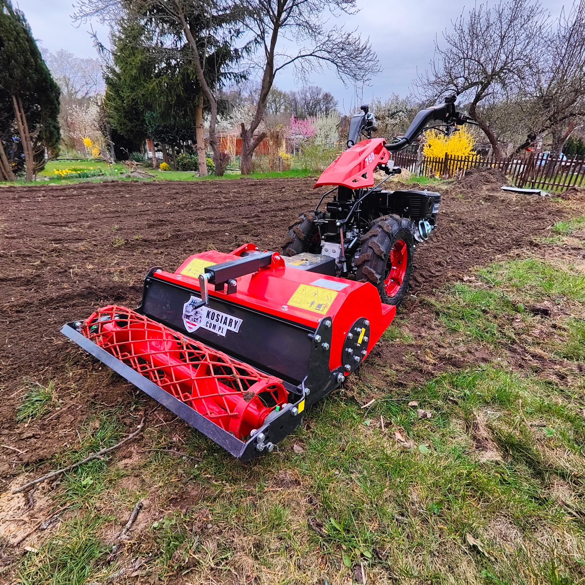 Czerwony glebogryz z logo Kosiary.com.pl na polu z przygotowaną ziemią, w tle kwitnące drzewa i drewniany płot.