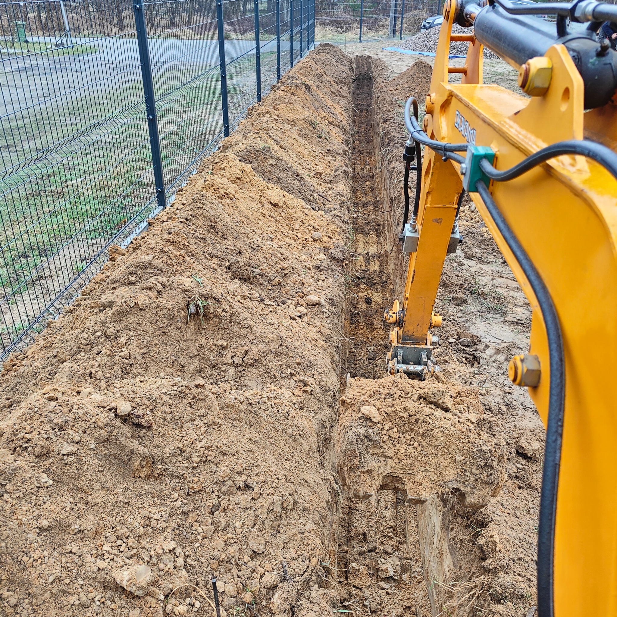 Wykopany wąski rów w ziemi wzdłuż ogrodzenia z siatki, wykonany żółtą koparką z widocznymi elementami hydraulicznymi.