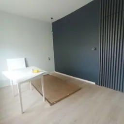 Pokój po malowaniu z ciemną ścianą i dekoracyjnymi panelami pionowymi. Na podłodze karton, a na stole akcesoria malarskie.