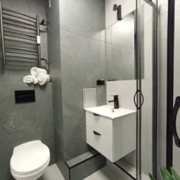 Nowoczesna łazienka w szarościach z białymi akcentami: umywalka z szafką, toaleta, grzejnik drabinkowy i kabina prysznicowa z czarnymi profilami. Minimalistyczny design.