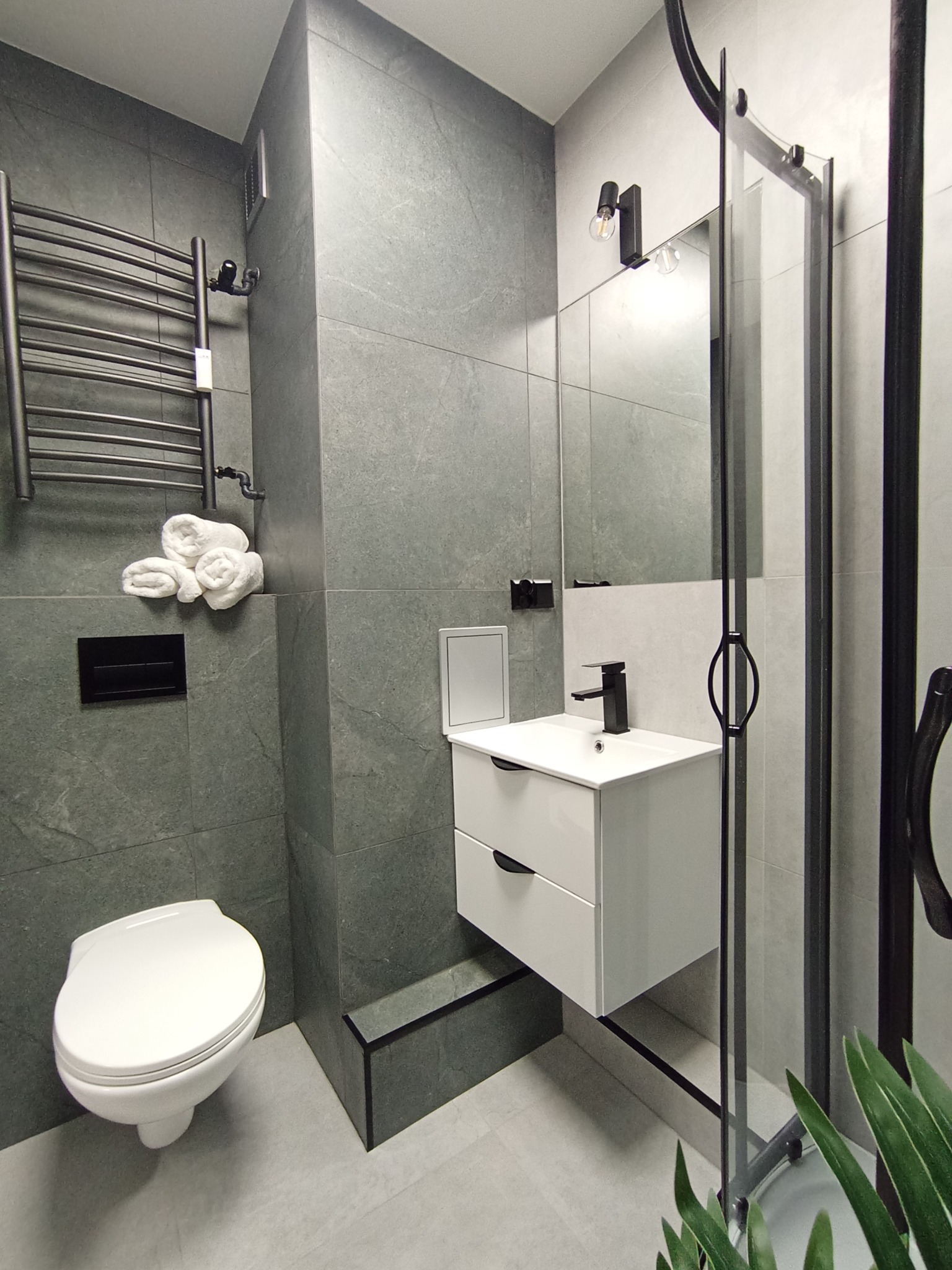 Nowoczesna łazienka w szarościach z białymi akcentami: umywalka z szafką, toaleta, grzejnik drabinkowy i kabina prysznicowa z czarnymi profilami. Minimalistyczny design.