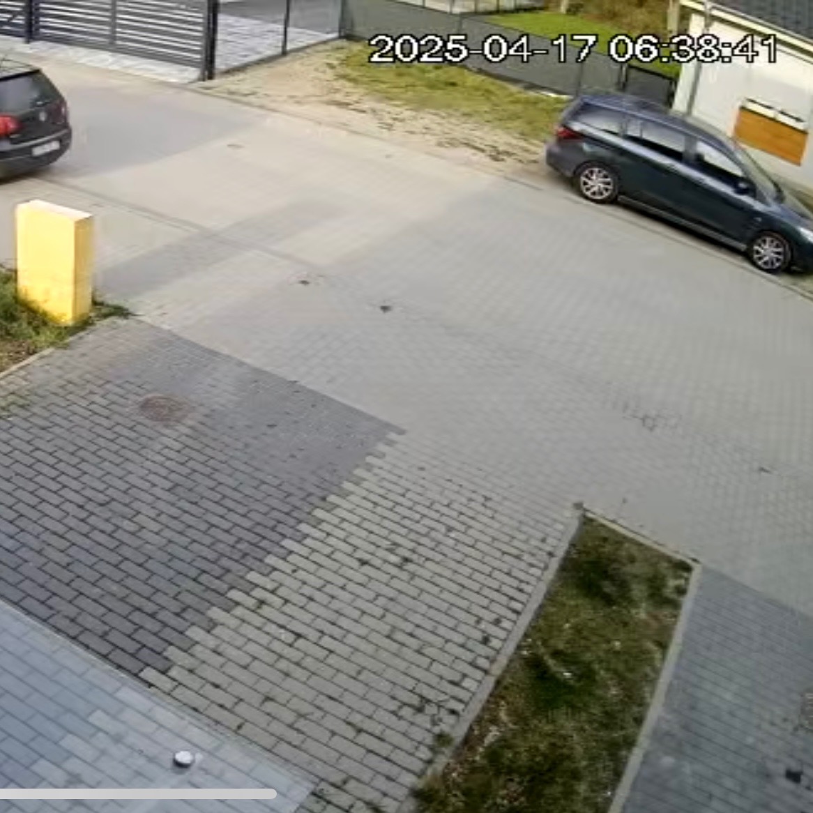 Obraz z kamery monitoringu: widok z góry na podjazd z kostki brukowej, dwa samochody zaparkowane po obu stronach, widoczna data i godzina na nagraniu.