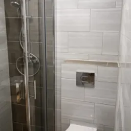 Nowoczesna łazienka z kabiną prysznicową ze srebrnymi ramami i deszczownicą oraz podwieszaną toaletą z chromowanym przyciskiem spłukującym, wykończona płytkami w odcieniach szarości i brązu.