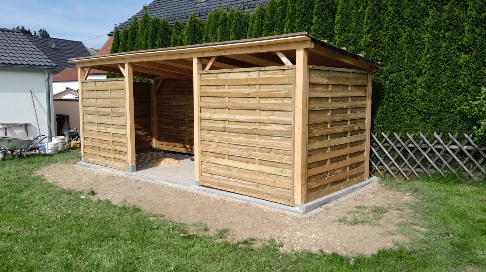 Drewniana wiata z ażurowymi ścianami, betonowa podstawa, widok z zewnątrz na tle zieleni.