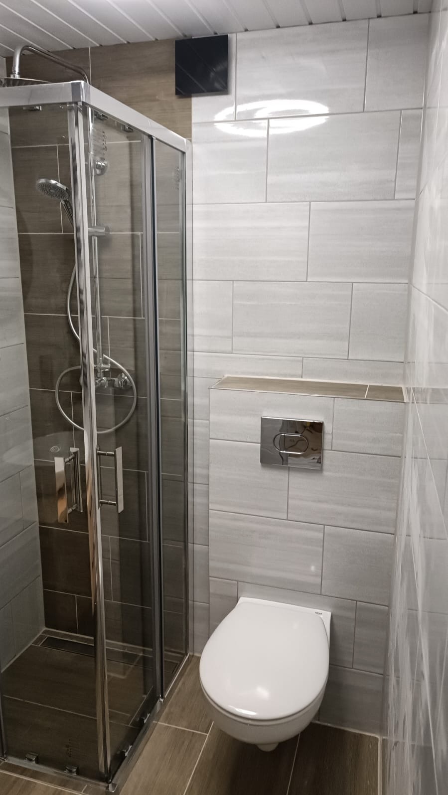 Nowoczesna łazienka z kabiną prysznicową ze srebrnymi ramami i deszczownicą oraz podwieszaną toaletą z chromowanym przyciskiem spłukującym, wykończona płytkami w odcieniach szarości i brązu.
