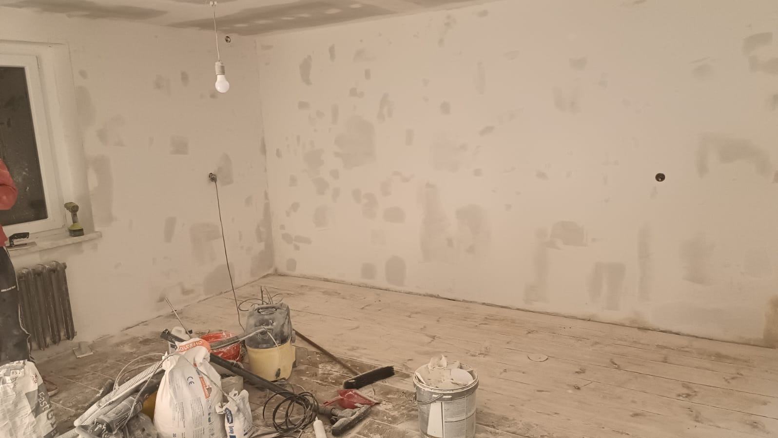 Pokój w trakcie prac przygotowawczych do malowania, ściany z widocznymi łatami szpachli, worki z gładzią i narzędzia na podłodze, surowa żarówka na kablu.