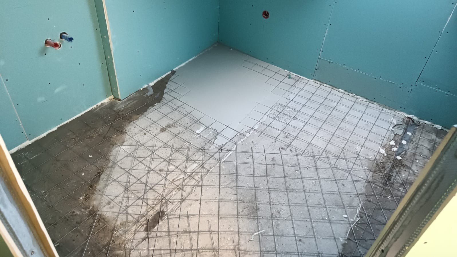 Przygotowanie podłoża pod posadzkę w łazience: wylewka częściowo pokrywa siatkę zbrojeniową, ściany wyłożone płytami kartonowo-gipsowymi, widoczne instalacje wodne.