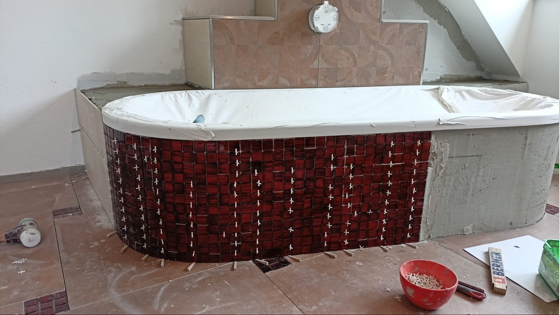Proces wykańczania wanny mozaiką z czerwonych płytek; widoczne krzyżyki dystansowe, poziomica, oraz fragmenty niedokończonej obudowy z surowego betonu.