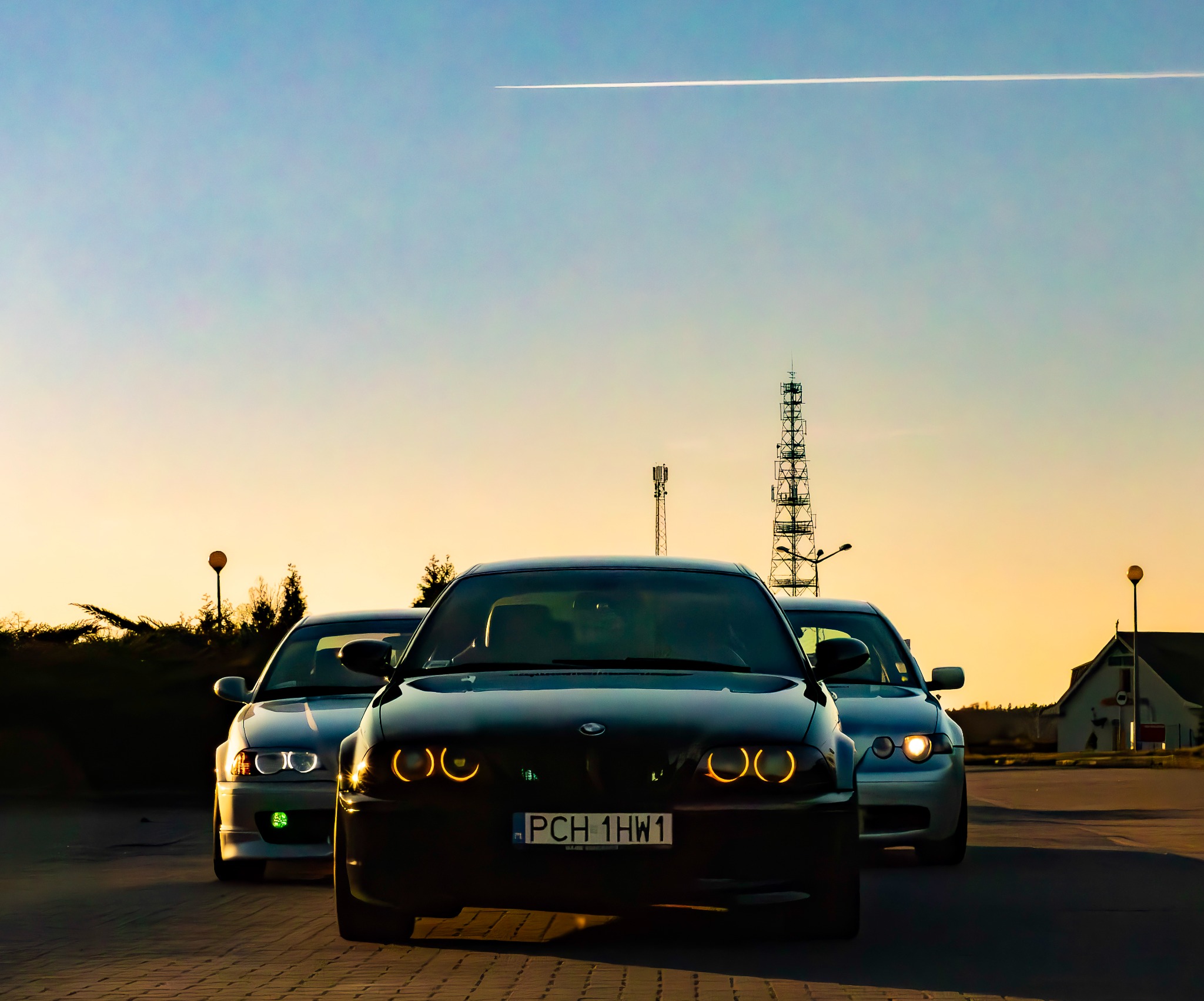 Trzy samochody BMW E46 ustawione w rzędzie, z włączonymi światłami, na tle zachodzącego słońca i wieży telekomunikacyjnej. Na niebie widoczny ślad po samolocie.