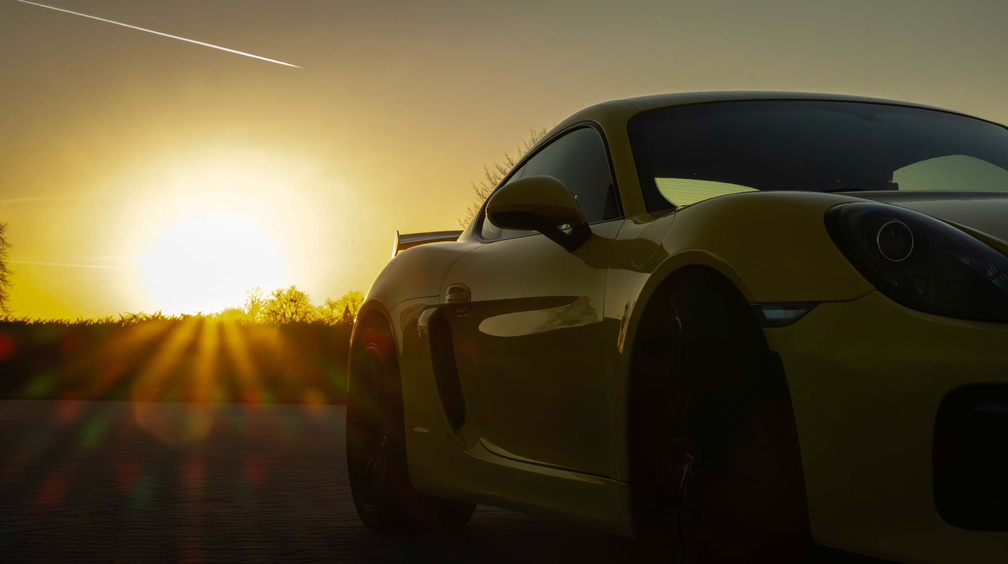 Żółty sportowy samochód Porsche Cayman w promieniach zachodzącego słońca, z widocznym śladem samolotu na niebie.