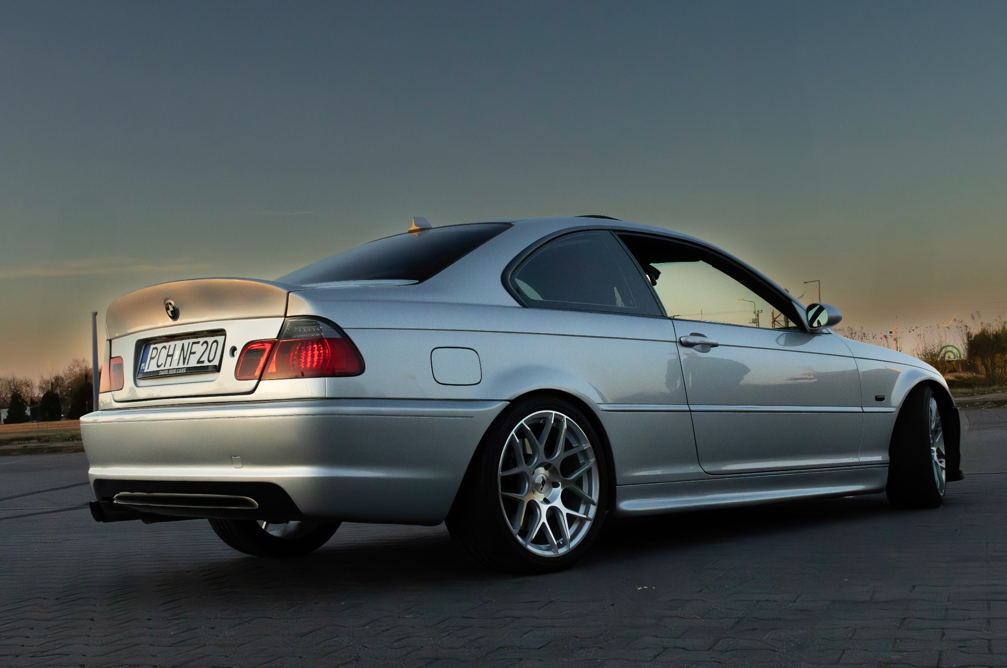 Srebrne BMW E46 Coupe z aluminiowymi felgami, widok z tyłu, tablica rejestracyjna PCH NF20, sfotografowane o zmierzchu na brukowanej powierzchni.