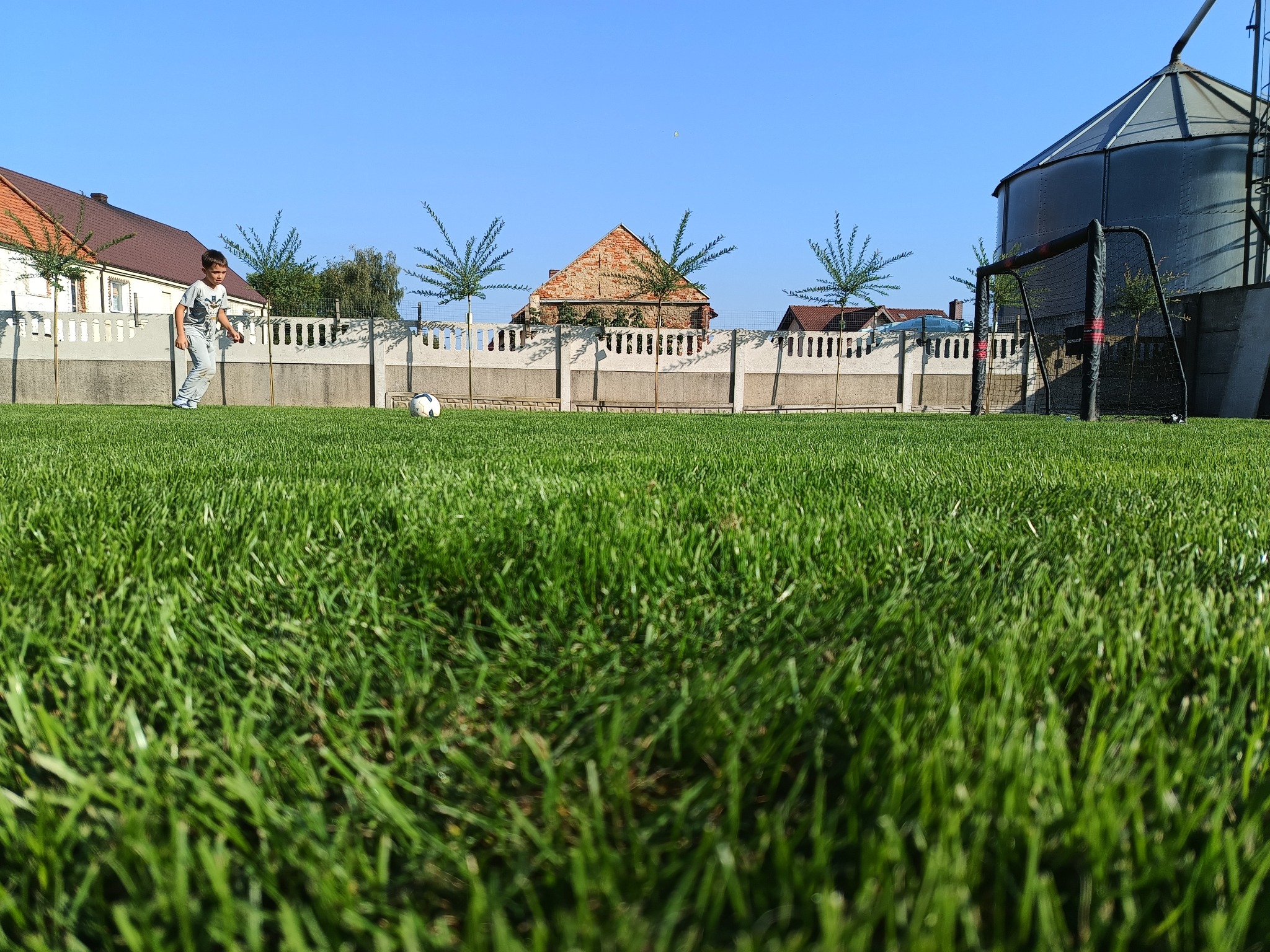 Chłopiec w szarej koszulce i spodniach kopie piłkę na intensywnie zielonym trawniku przed ogrodzeniem z młodymi drzewkami, w tle budynki i silos.
