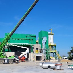 PROI Damian Borek - Budowa hali: zielona konstrukcja stalowa w trakcie montażu, widoczny dźwig, elementy instalacji i pracownicy w kaskach. Błękitne niebo w tle.