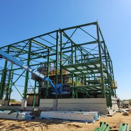 PROI Damian Borek - Montaż konstrukcji stalowej hali z użyciem podnośnika Genie Z-80/60. Widoczny fragment budynku i silos. Słoneczny dzień, błękitne niebo.