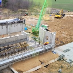 PROI Damian Borek - Budowa fundamentów z betonu: widok z góry na plac budowy z dźwigiem, pracownikami w kaskach i maszynami budowlanymi. Etap wylewania fundamentów.