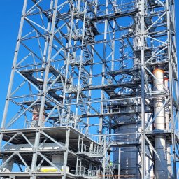 PROI Damian Borek - Konstrukcja stalowa hali przemysłowej w trakcie budowy, widok z dołu na szkielet z licznymi schodami i pomostami, na tle błękitnego nieba. Na dole widoczne podnośniki.