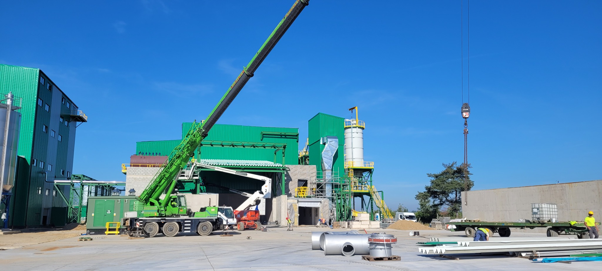 Budowa hali: zielona konstrukcja stalowa w trakcie montażu, widoczny dźwig, elementy instalacji i pracownicy w kaskach. Błękitne niebo w tle.