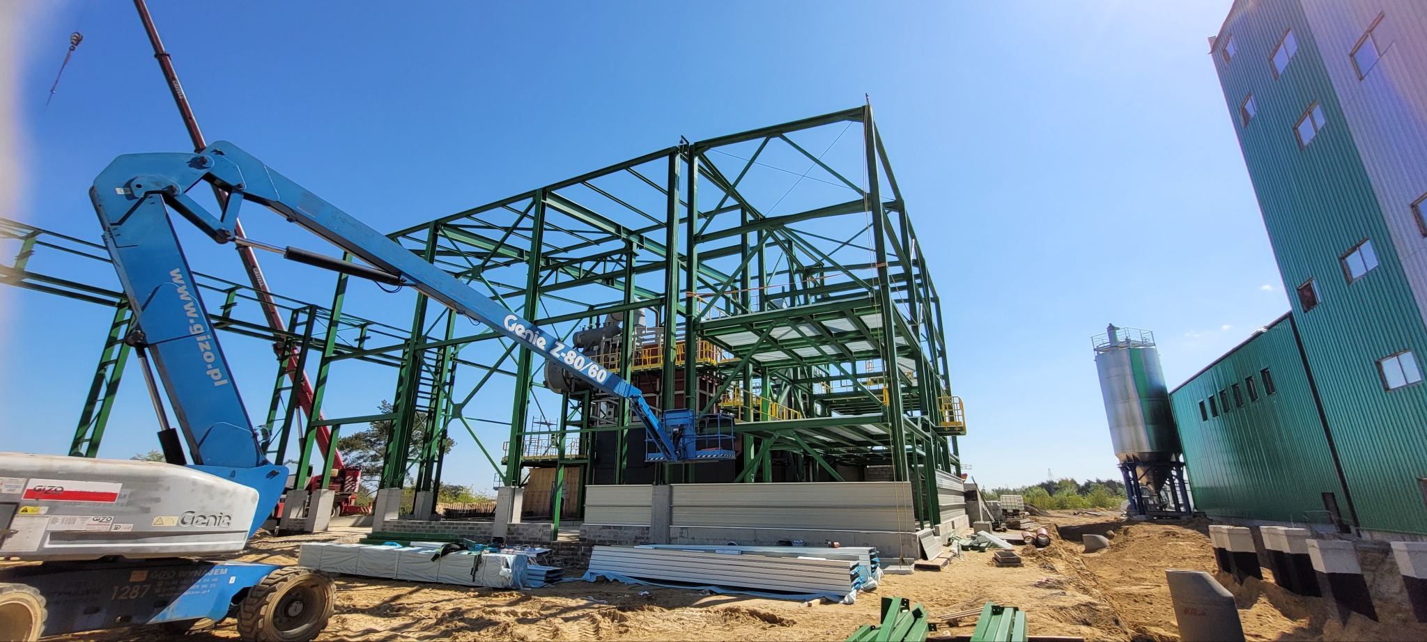 Montaż konstrukcji stalowej hali z użyciem podnośnika Genie Z-80/60. Widoczny fragment budynku i silos. Słoneczny dzień, błękitne niebo.