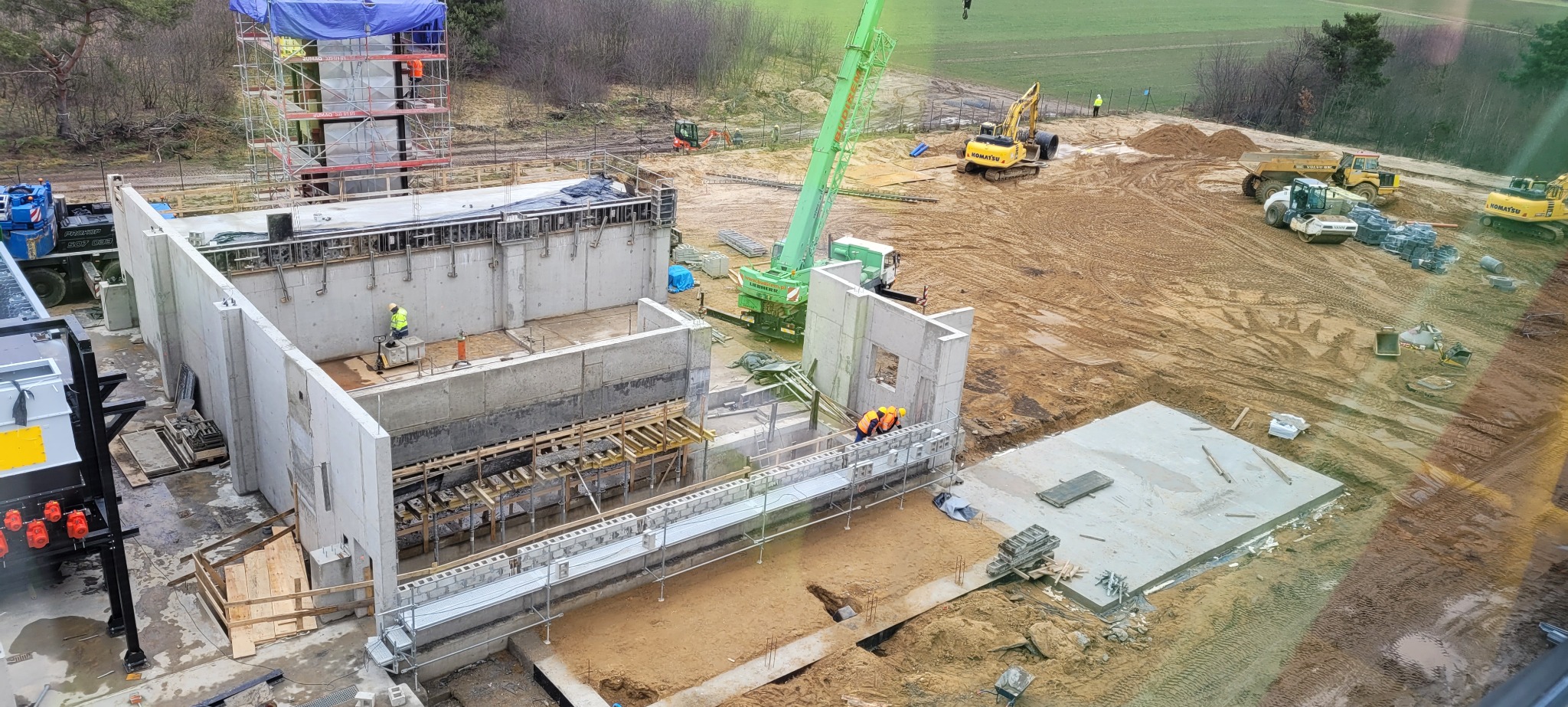 Budowa fundamentów z betonu: widok z góry na plac budowy z dźwigiem, pracownikami w kaskach i maszynami budowlanymi. Etap wylewania fundamentów.