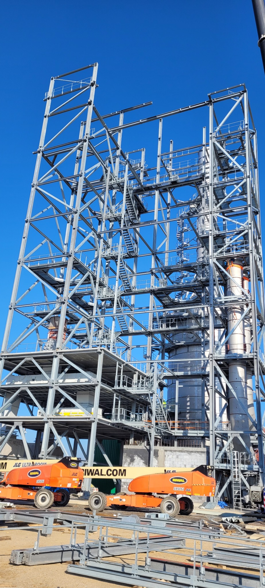 Konstrukcja stalowa hali przemysłowej w trakcie budowy, widok z dołu na szkielet z licznymi schodami i pomostami, na tle błękitnego nieba. Na dole widoczne podnośniki.