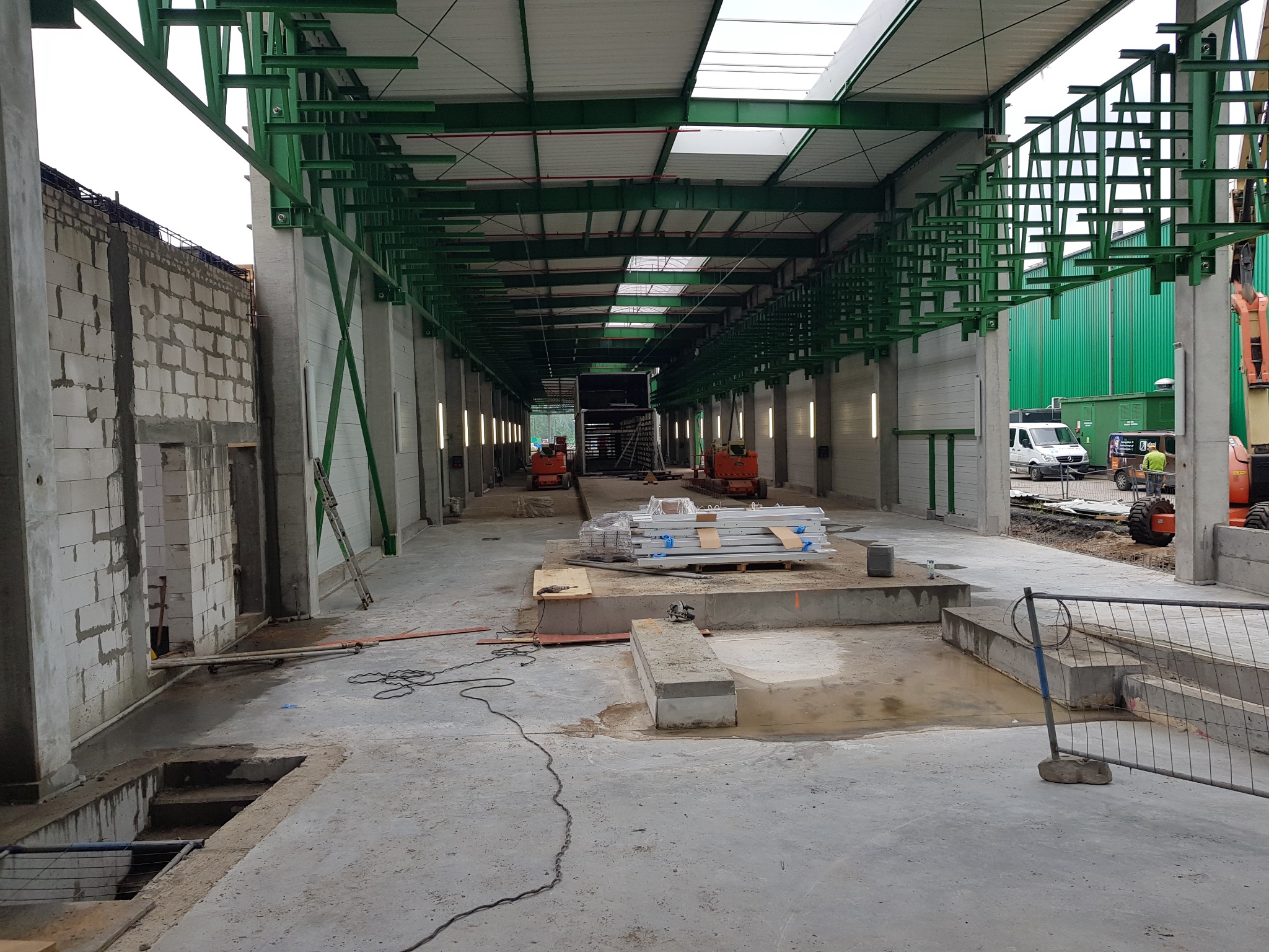 Wnętrze hali w trakcie budowy: surowy beton, zielona konstrukcja stalowa pod dachem, materiały budowlane, częściowo wymurowane ściany. Widoczne maszyny budowlane na zewnątrz.