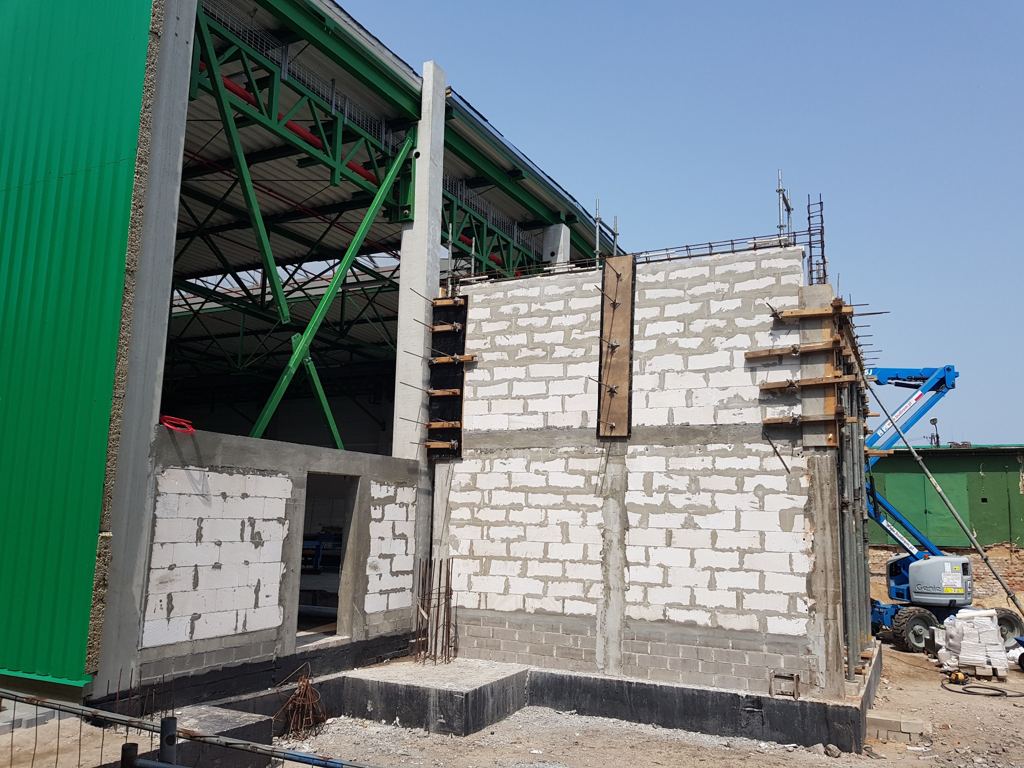 Wznoszenie ścian budynku z betonu komórkowego Ytong, widoczne szalunki i zbrojenia. W tle konstrukcja stalowa hali i podnośnik koszowy.