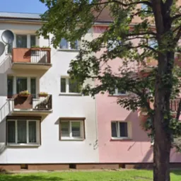 Dwupiętrowy budynek z balkonami, z anteną satelitarną na jednym z nich. Część budynku w kolorze białym, część w odcieniach różu. Duże drzewo z przodu.