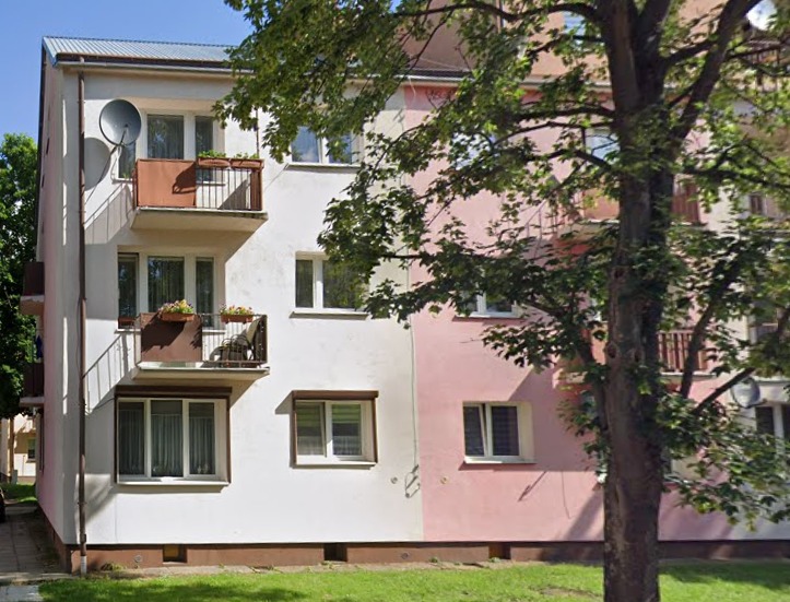 Dwupiętrowy budynek z balkonami, z anteną satelitarną na jednym z nich. Część budynku w kolorze białym, część w odcieniach różu. Duże drzewo z przodu.