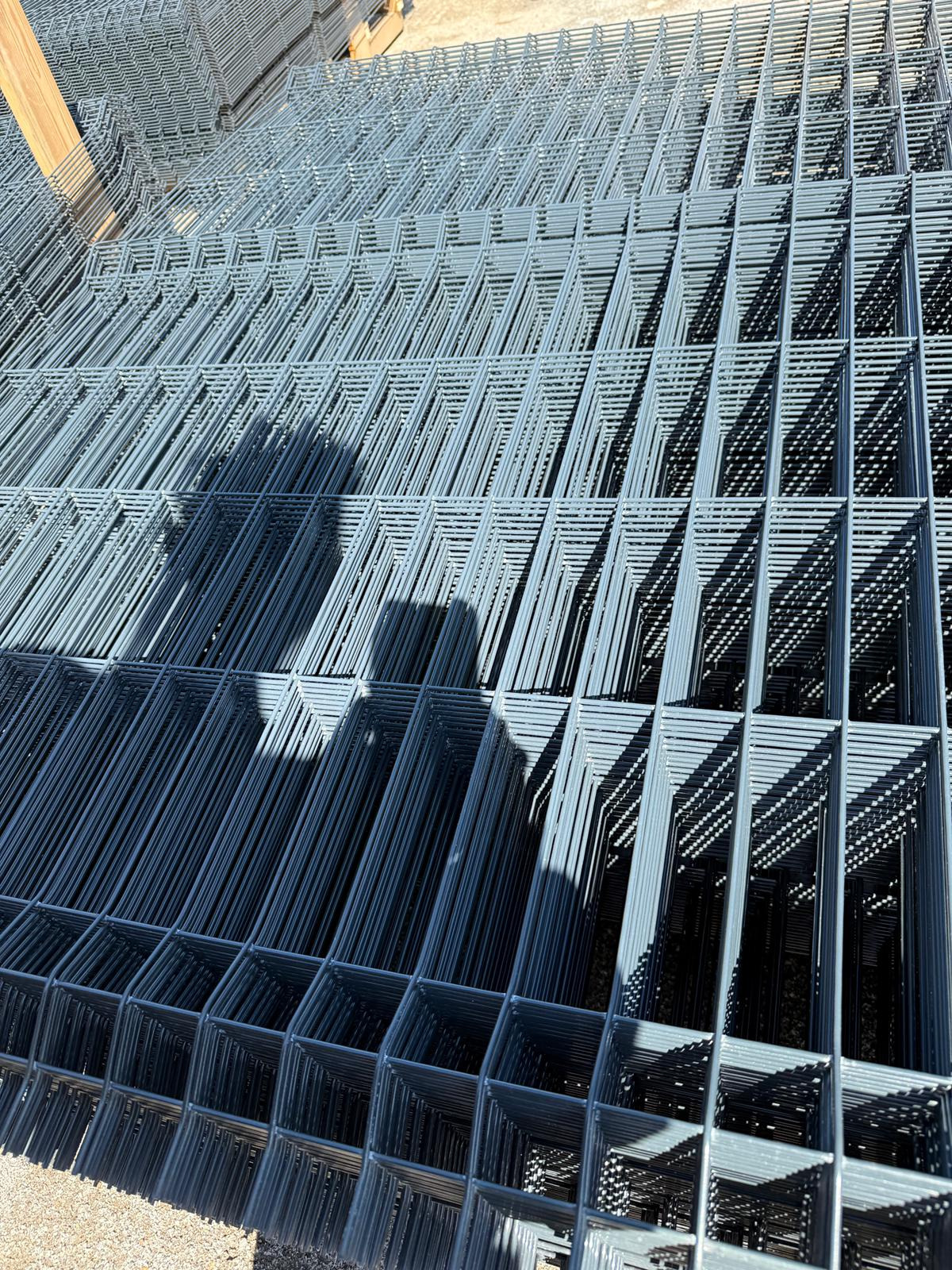 Stos paneli ogrodzeniowych z metalu, przygotowanych do montażu w Piotrowicach. Widoczne regularne oczka siatki i cienie rzucane przez elementy konstrukcyjne.