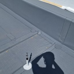 Podgórscy z wami od lat - Szary, płaski dach pokryty papą termozgrzewalną z widocznym mocowaniem i cieniem osoby, ukazujący detale hydroizolacji i zabezpieczeń.