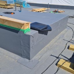 Podgórscy z wami od lat - Szary, płaski dach pokryty papą termozgrzewalną z detalami obróbek blacharskich, drewnianymi elementami i sprzętem dekarskim w tle. Widoczne materiały izolacyjne i palety.