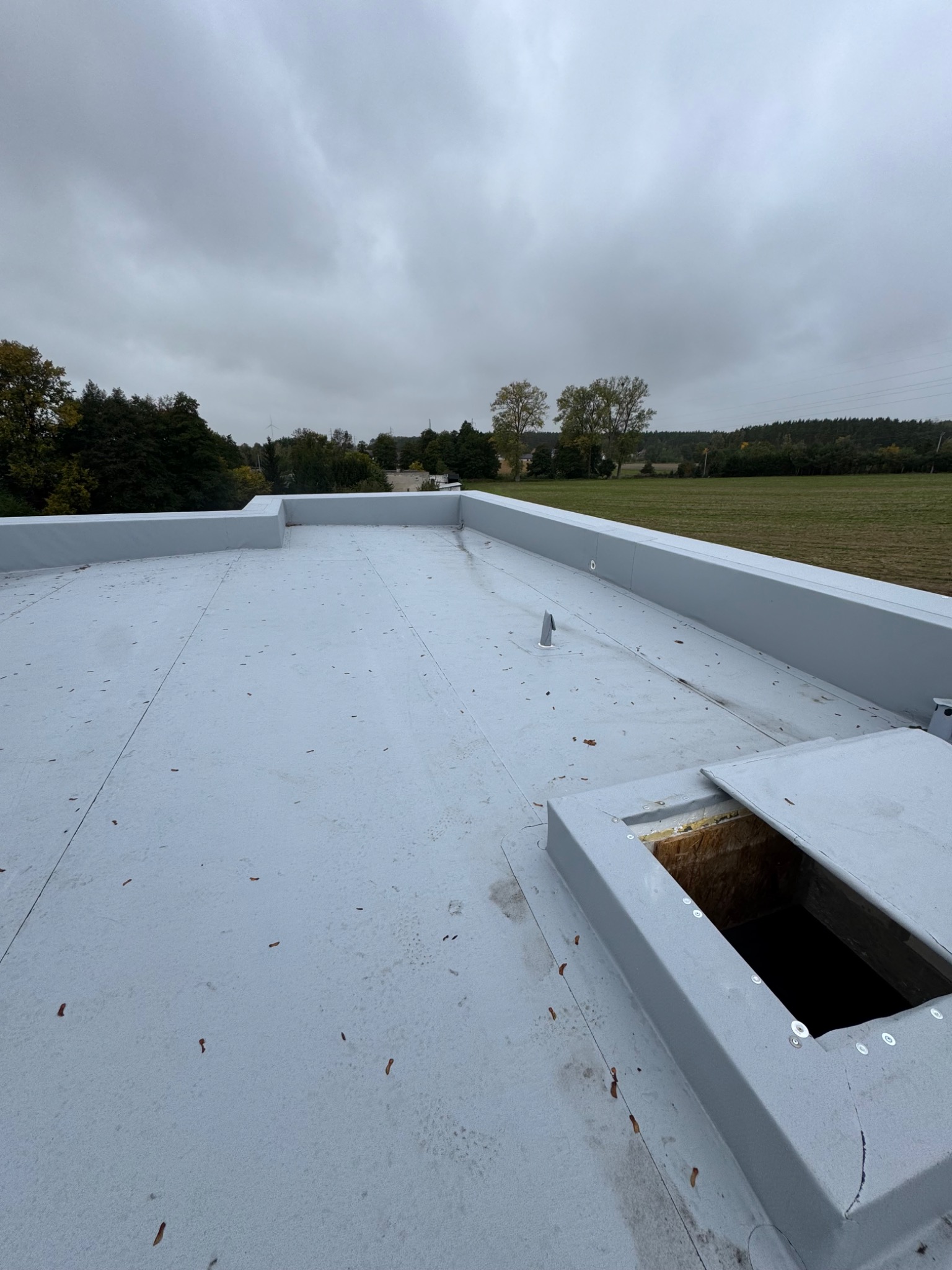 Szary, płaski dach z otwartym włazem, widok z góry. W tle drzewa i pochmurne niebo. Widoczne ślady po liściach na powierzchni dachu.