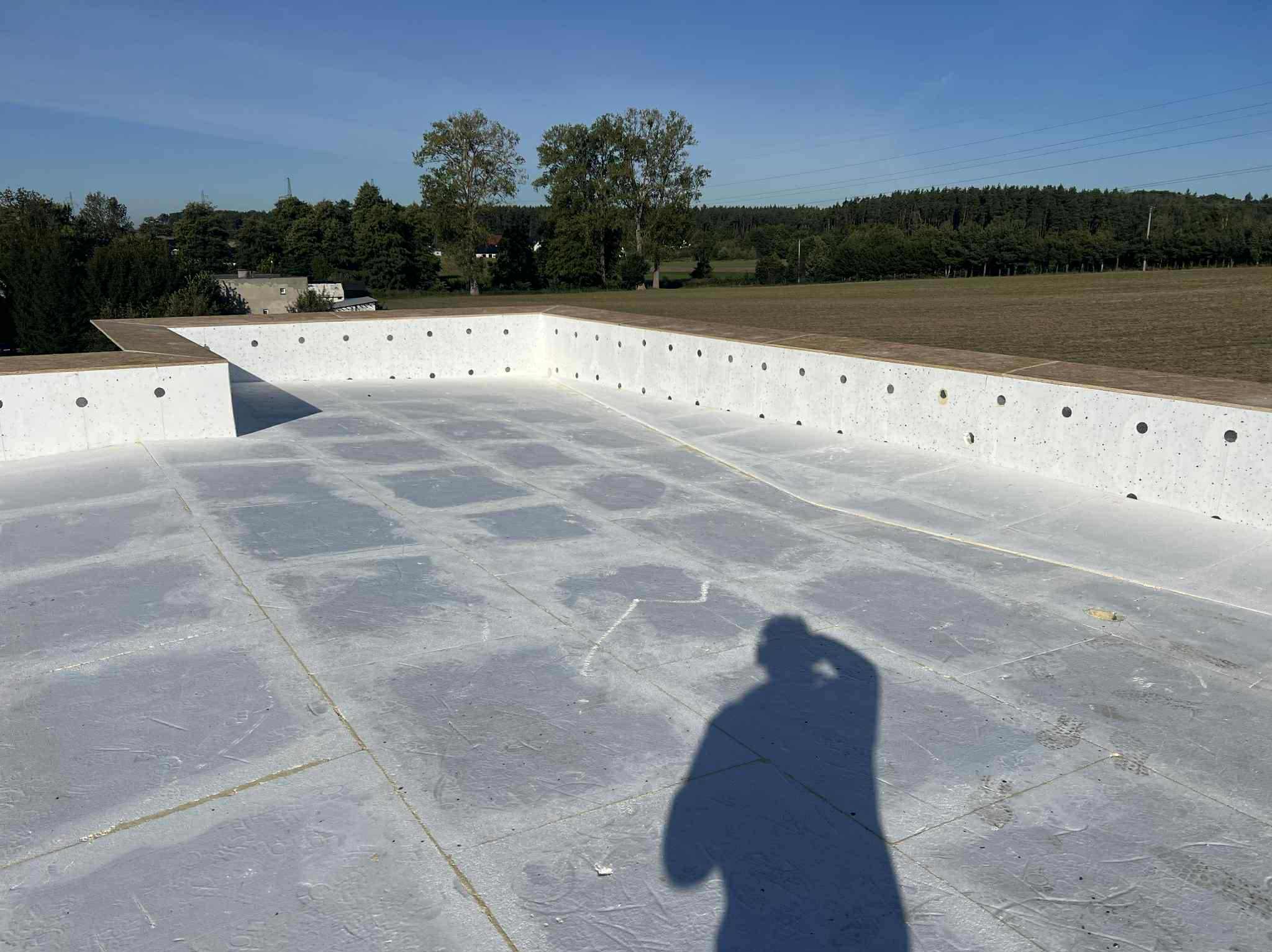 Biała, płaska powierzchnia dachu z otworami, otoczona niską ścianką. W tle drzewa i niebo. Cień osoby w prawym dolnym rogu. Widok z góry.