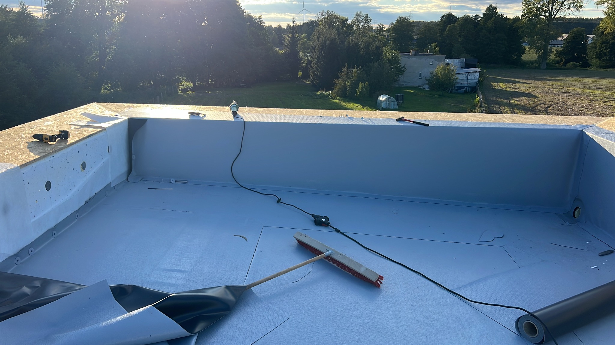 Szary dach płaski z narzędziami i rolką papy w słoneczny dzień. Widoczne detale izolacji i obróbki blacharskiej. W tle zieleń drzew i budynki.