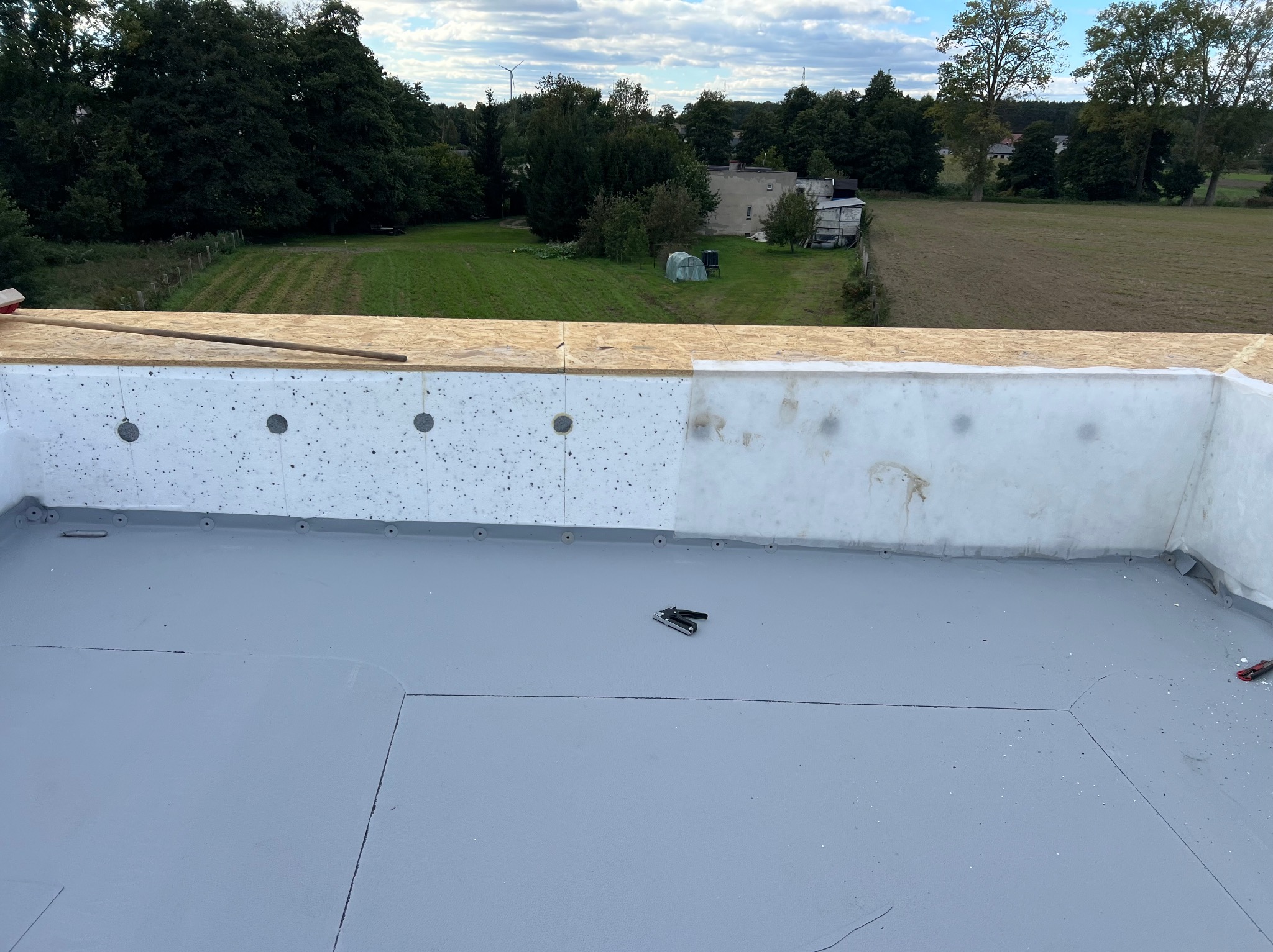 Szary, płaski dach z izolacją i narzędziami, widok z góry na tle zieleni. Elementy drewniane i biała izolacja. Prace dekarskie w toku, etap izolacji i pokrycia.