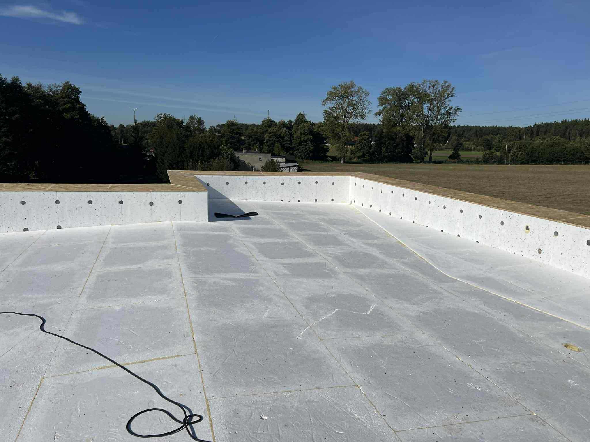 Płaski dach pokryty szarą membraną hydroizolacyjną z widocznymi łączeniami płyt, obrzeża z białego betonu z otworami, w tle zieleń i błękitne niebo.