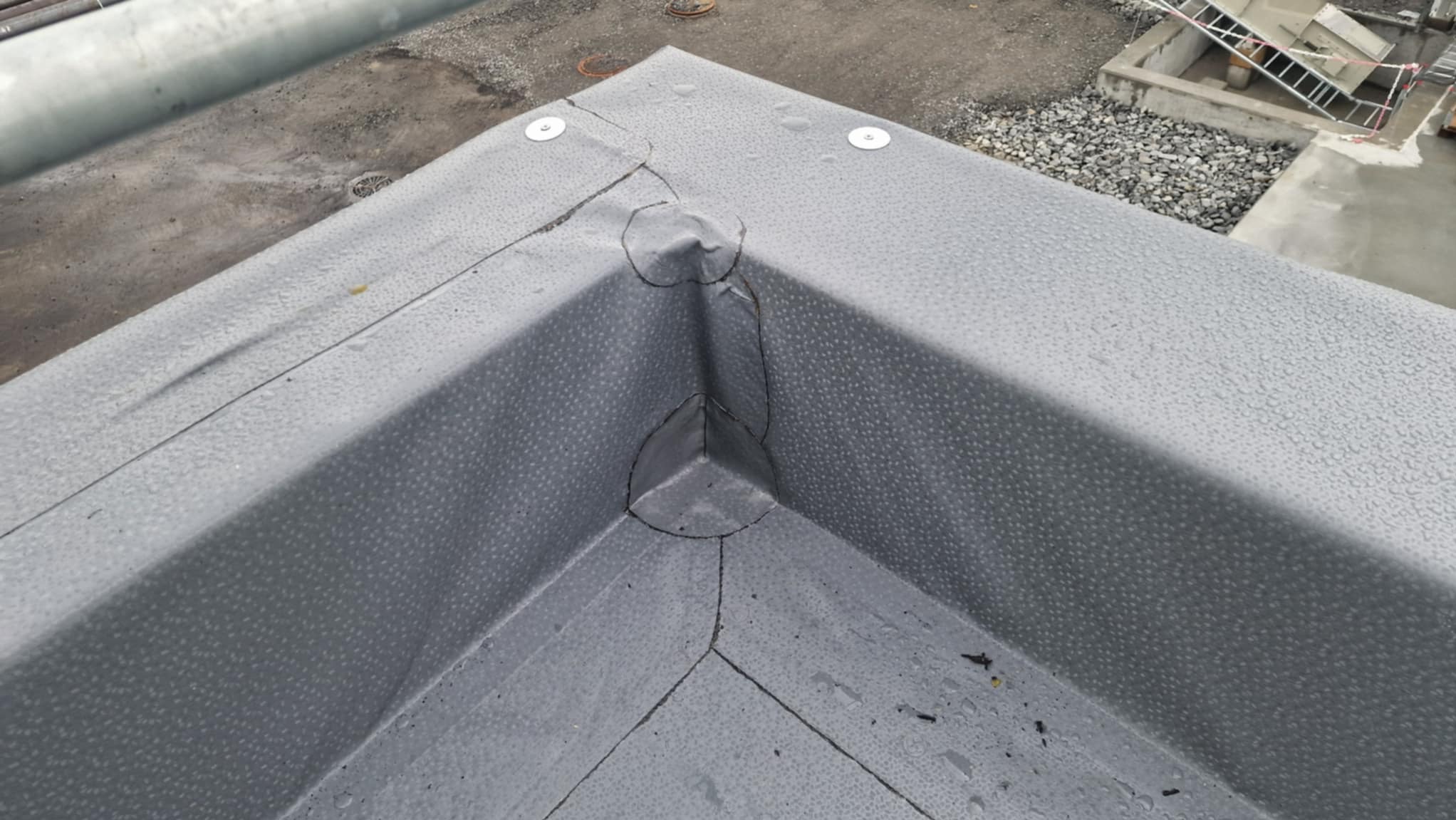 Szczegółowe ujęcie narożnika dachu pokrytego szarą membraną z widocznymi łączeniami i elementami mocującymi. W tle widoczny fragment dachu i drabina.