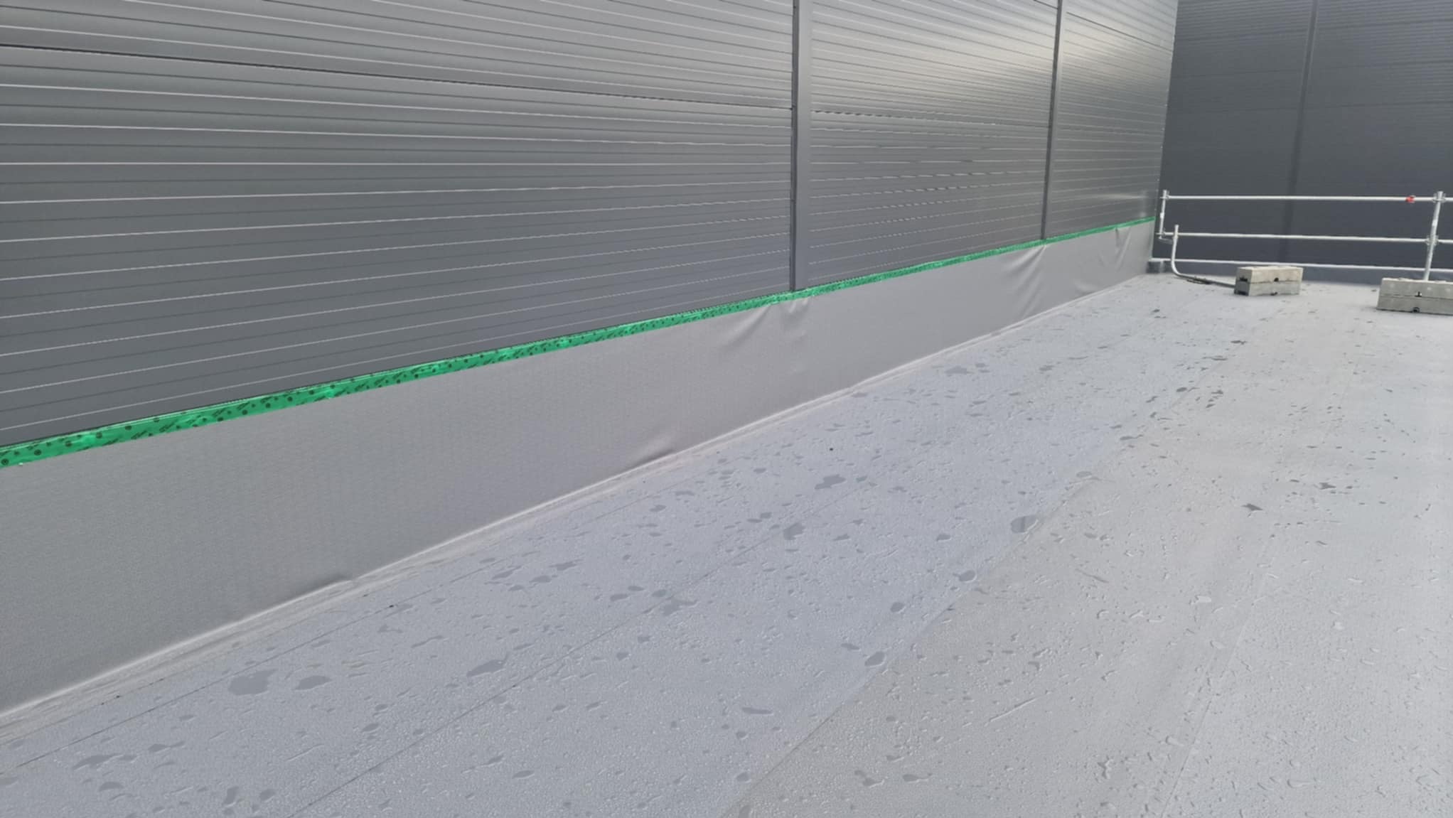 Szara, płaska powierzchnia dachu przemysłowego z widoczną izolacją pionową przy ścianie, zabezpieczoną zieloną taśmą. W tle ściana z blachy trapezowej.