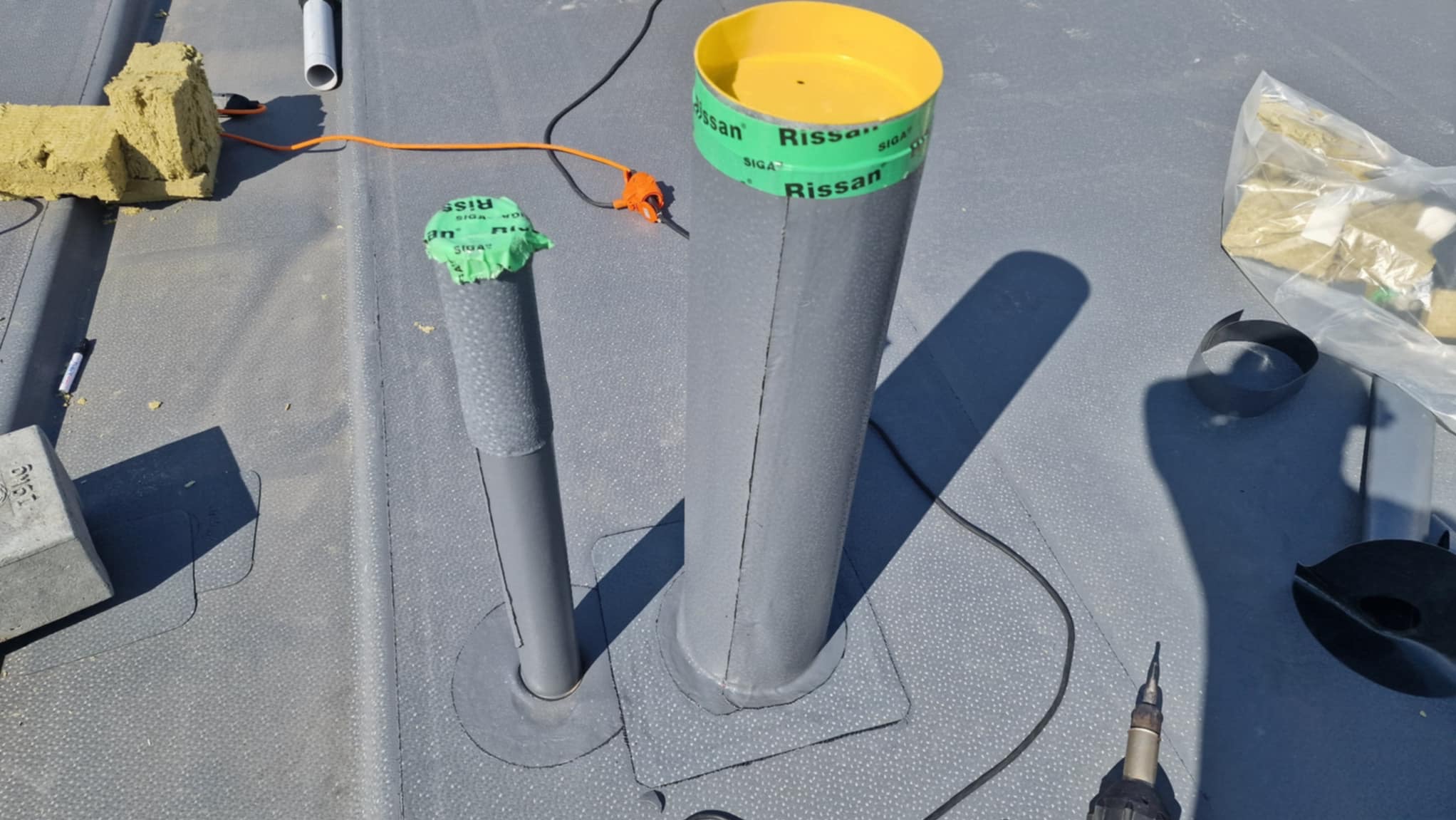 Szary dach z papy z dwoma kominami wentylacyjnymi uszczelnionymi taśmą Siga Rissan, widoczne narzędzia i materiały izolacyjne. Prace dekarskie w toku.