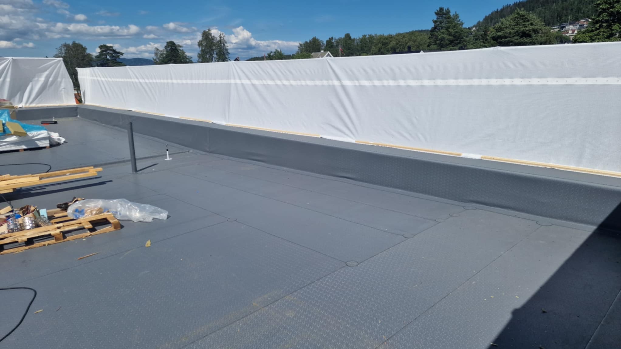 Szary dach płaski z widoczną membraną i zabezpieczeniem z białej plandeki. Na dachu leżą palety z materiałami budowlanymi. W tle drzewa i błękitne niebo.