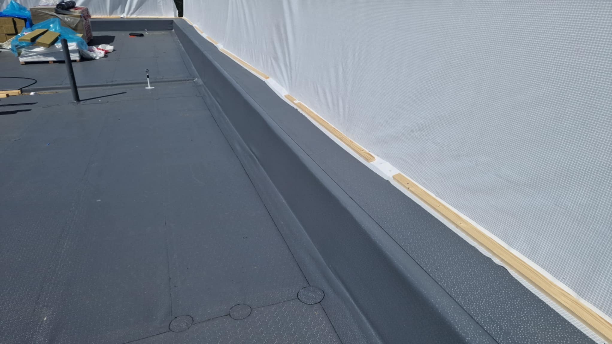 Szary, płaski dach z widoczną membraną i drewnianymi elementami wykończeniowymi przy krawędzi. Widoczne materiały budowlane w tle. Ujęcie z góry, podkreślające detale.