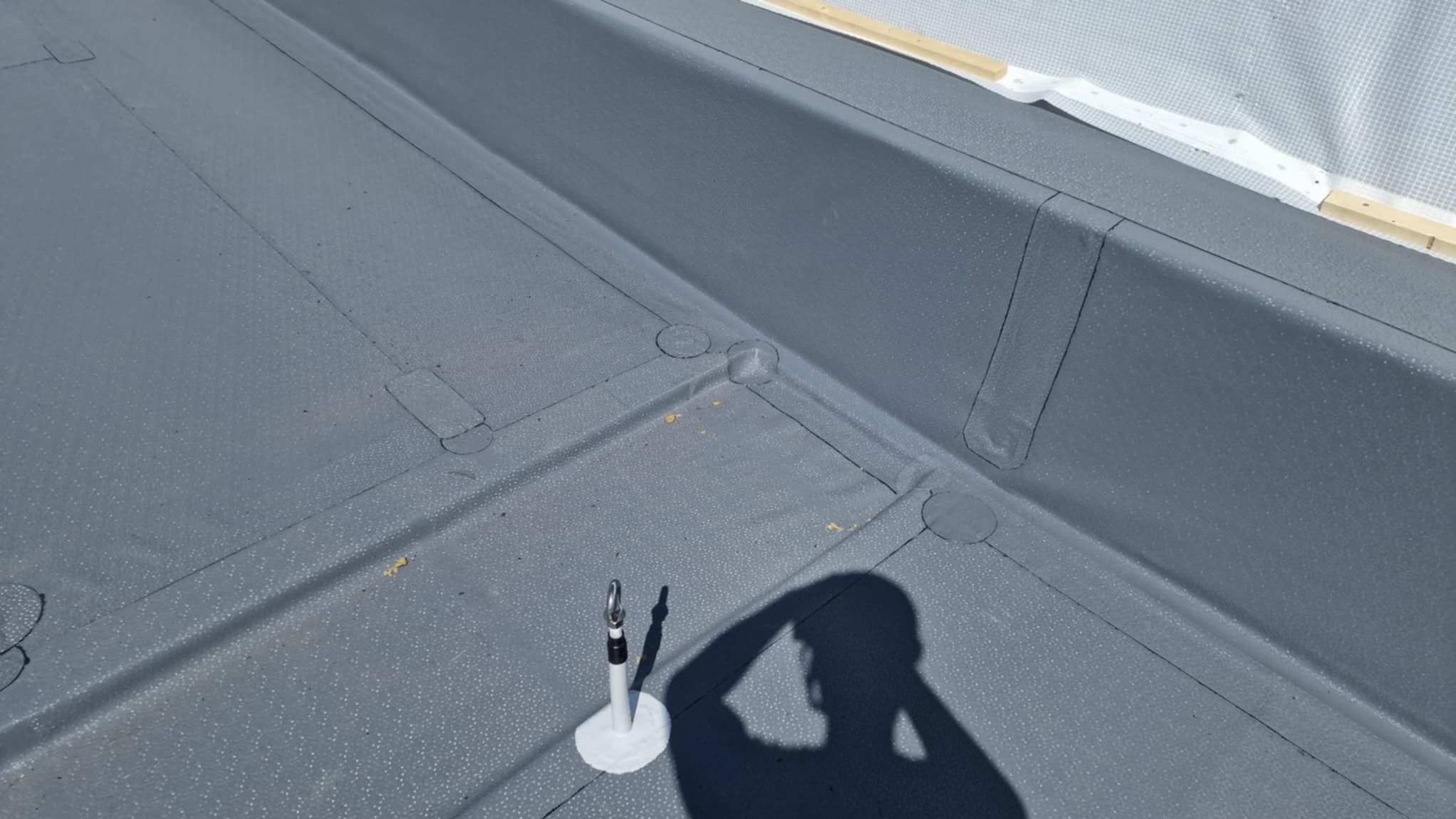 Szary, płaski dach pokryty papą termozgrzewalną z widocznym mocowaniem i cieniem osoby, ukazujący detale hydroizolacji i zabezpieczeń.