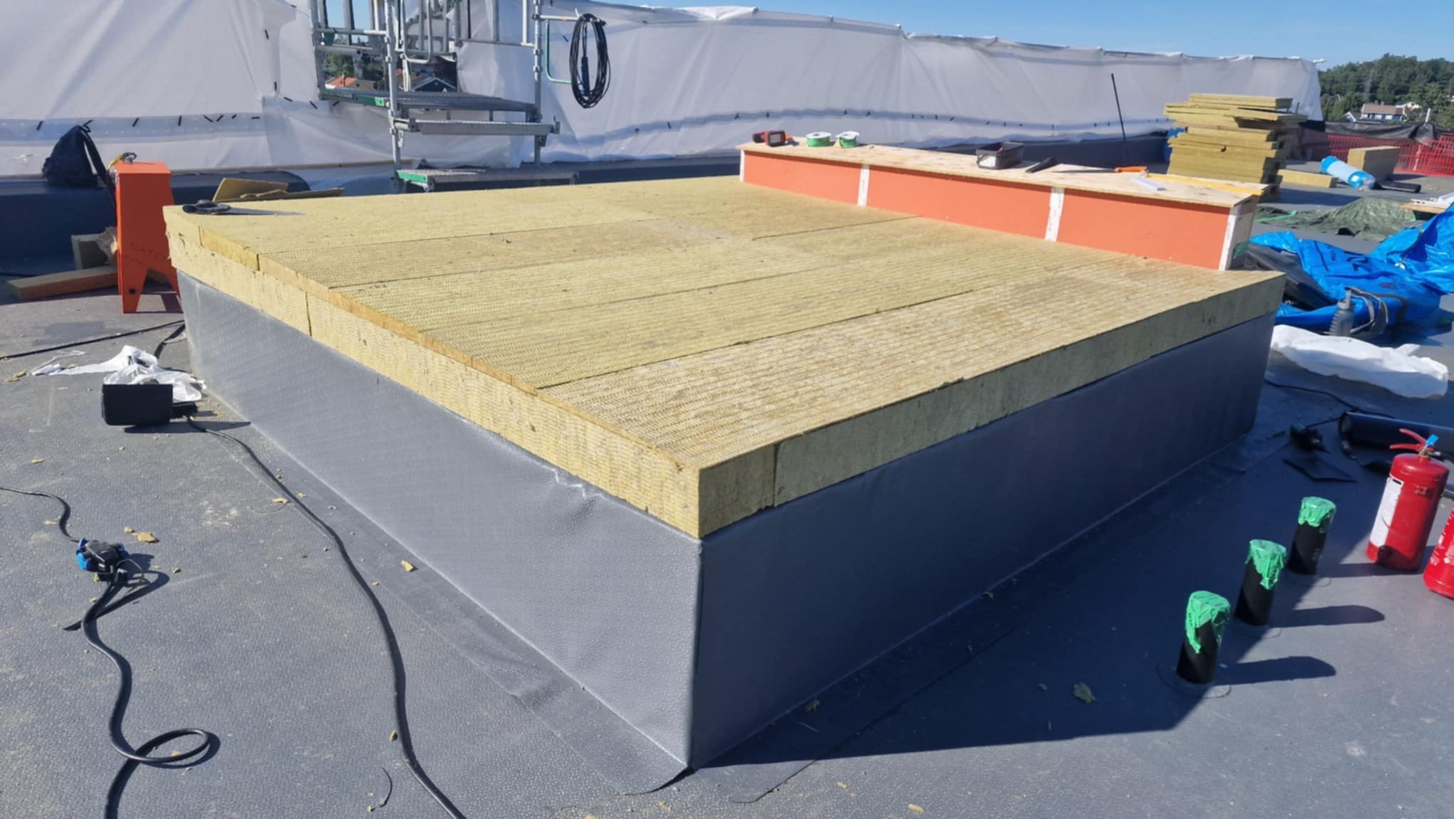 Izolacja termiczna dachu płaskiego z wełny mineralnej, zabezpieczona szarą membraną. Widoczne materiały budowlane i narzędzia na tle jasnego nieba.