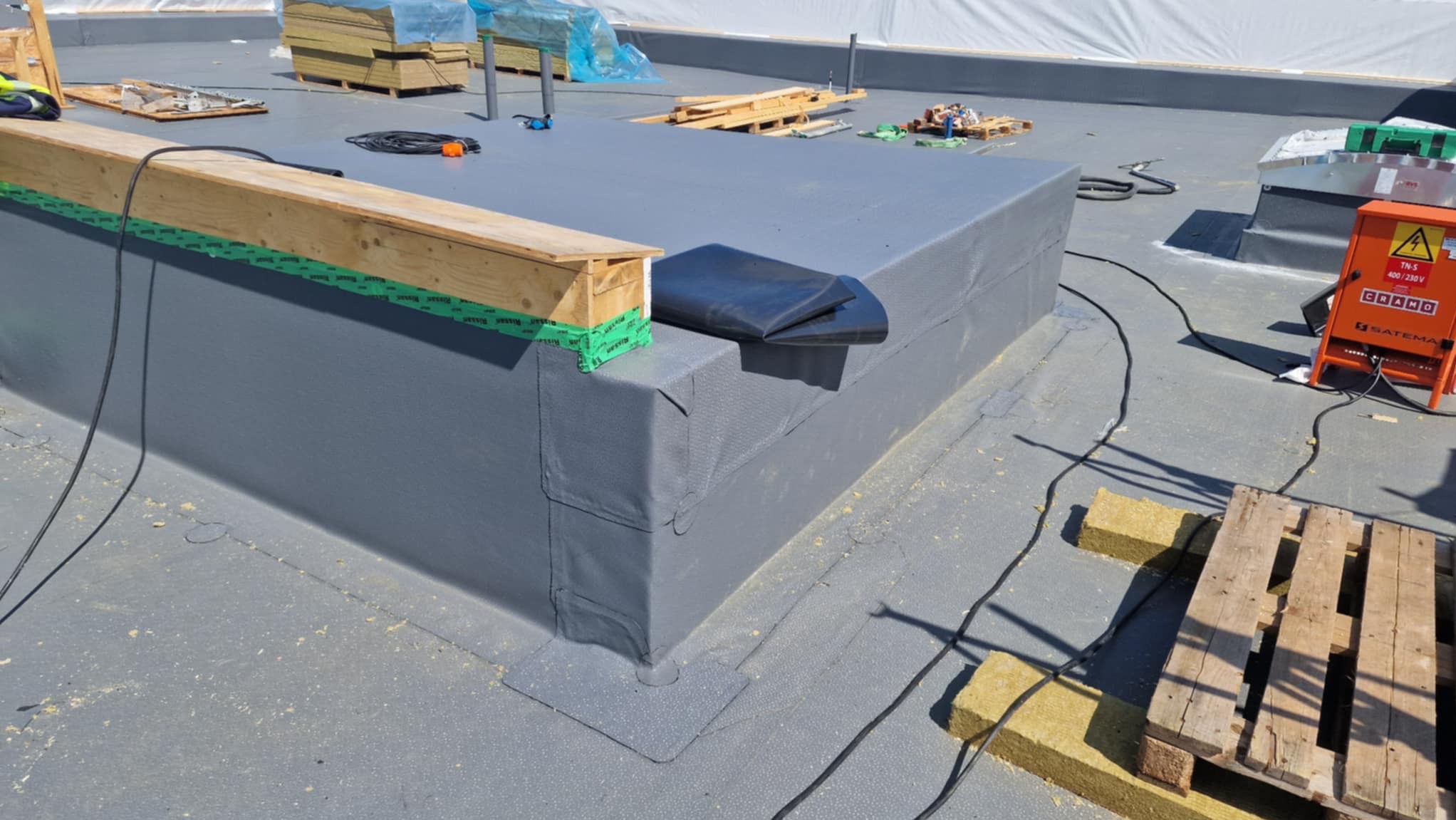 Szary, płaski dach pokryty papą termozgrzewalną z detalami obróbek blacharskich, drewnianymi elementami i sprzętem dekarskim w tle. Widoczne materiały izolacyjne i palety.